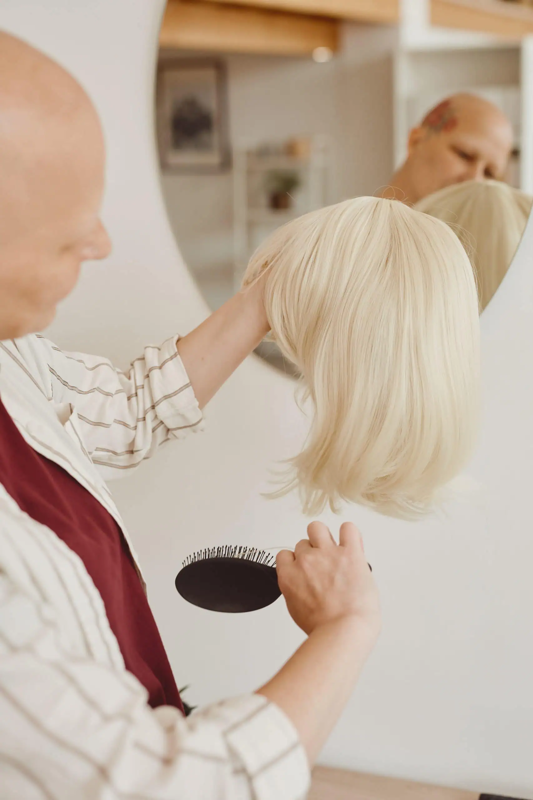 Bald Woman Caring for Wig Bald woman brushing wig while standing by mirror at home.